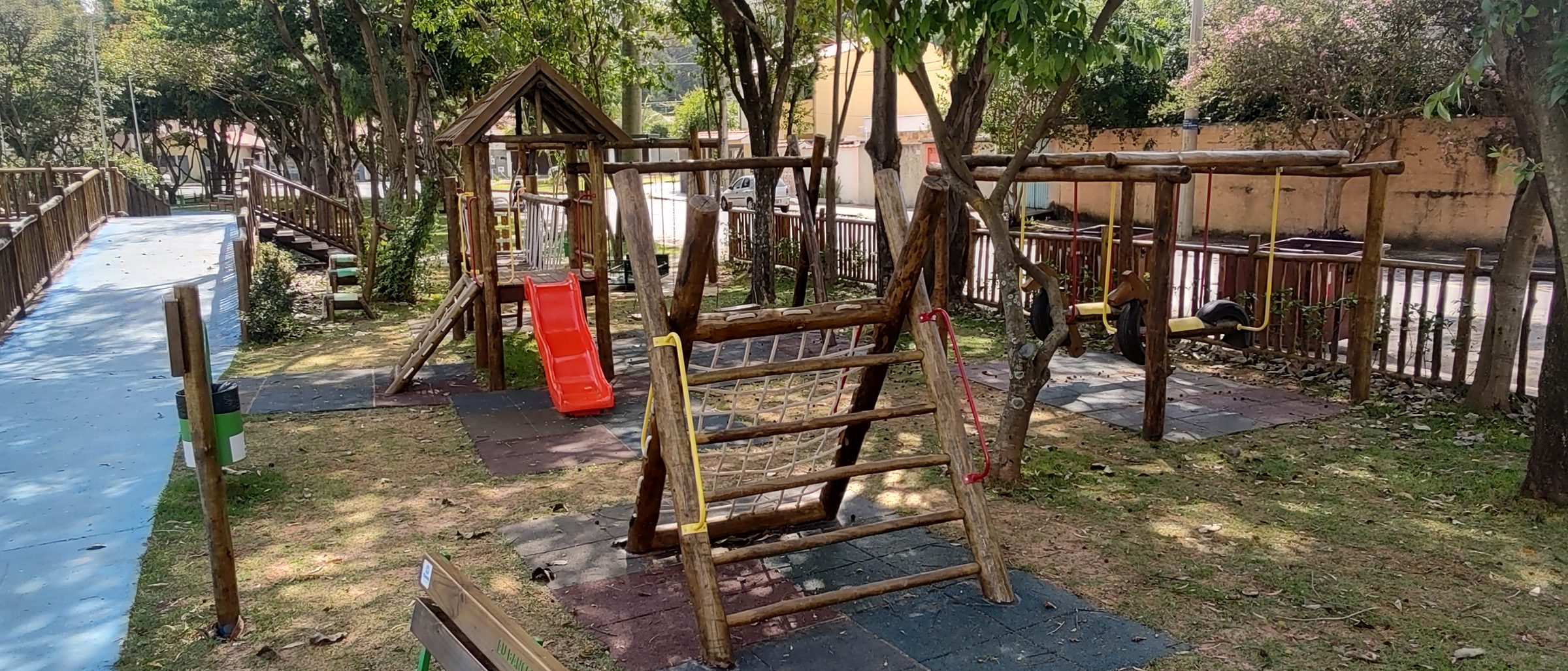 Conjunto de playground em toras de eucalipto com escorregador vermelho, barras amarelas e trepa-trepa sobre piso emborrachado, à sombra de árvores, com banco de madeira em primeiro plano