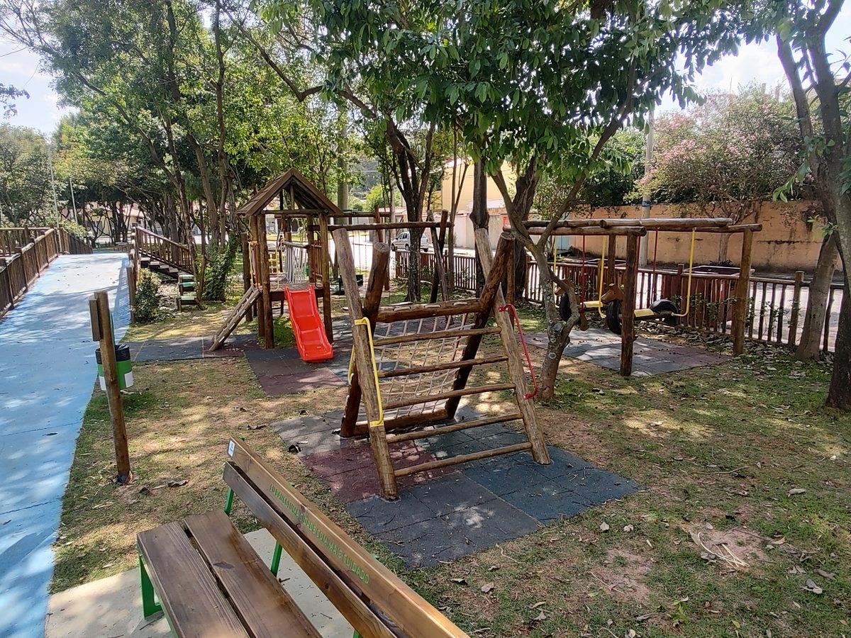 Conjunto de playground em toras de eucalipto com escorregador vermelho, barras amarelas e trepa-trepa sobre piso emborrachado, à sombra de árvores, com banco de madeira em primeiro plano