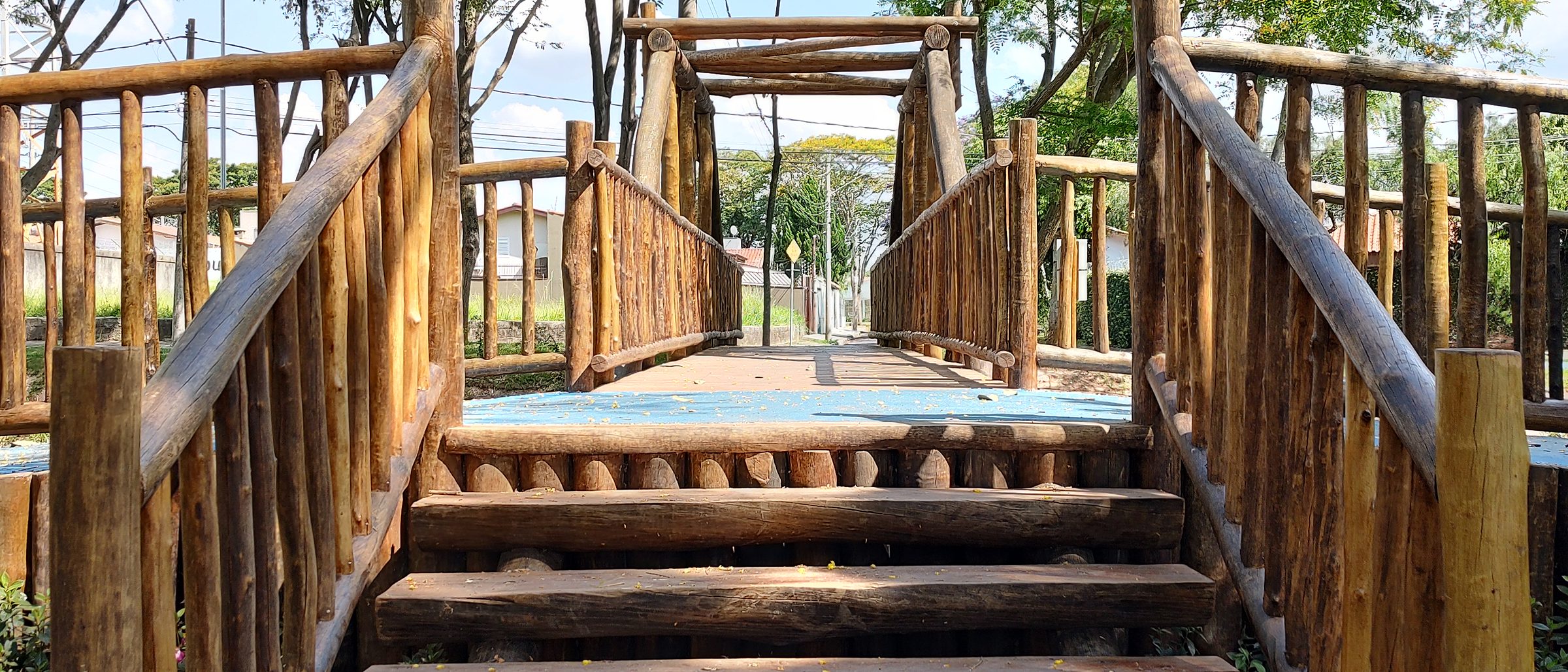 Estrutura elevada de toras de eucalipto com rampa de degraus e guarda-corpo de madeira rústica, sobre piso de terra com folhas secas, entre árvores