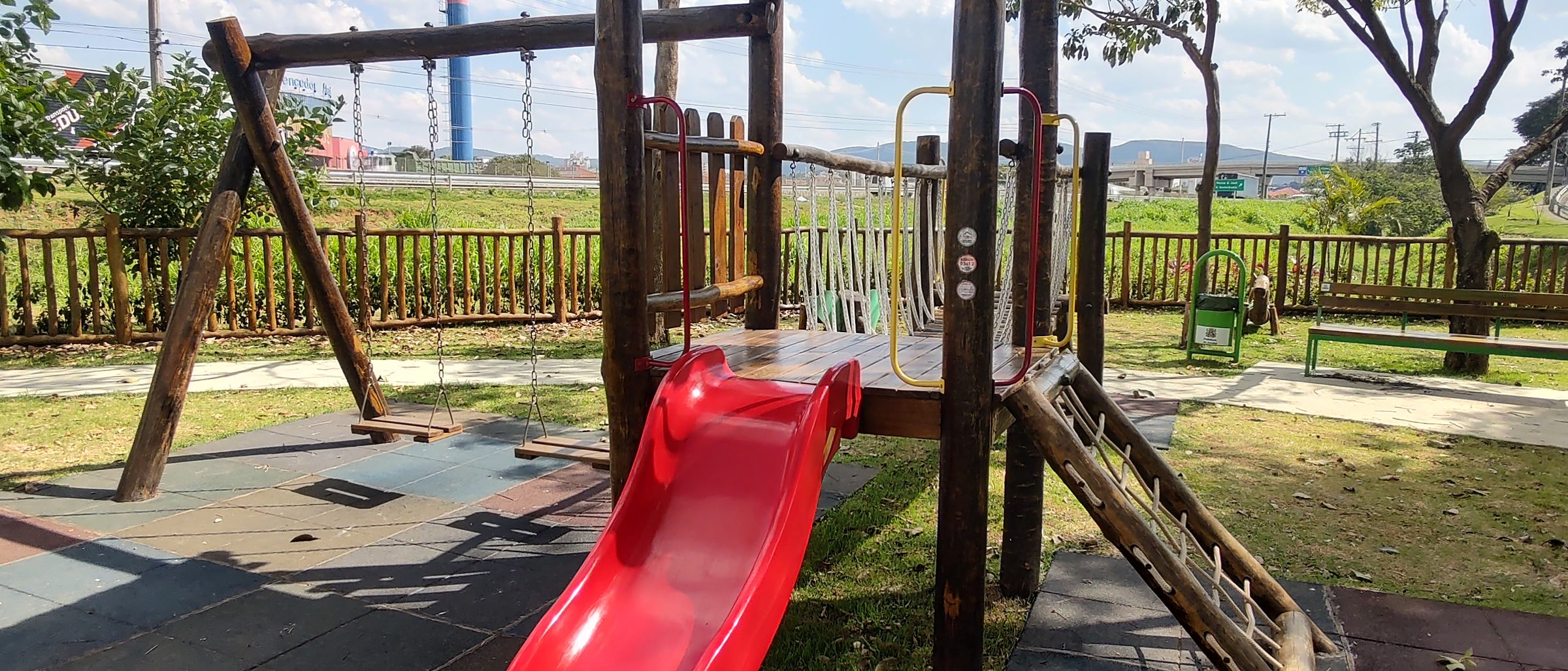 Torre de playground em toras de eucalipto com telhado, escorregador vermelho, balanço e piso de placas de borracha em praça de condomínio
