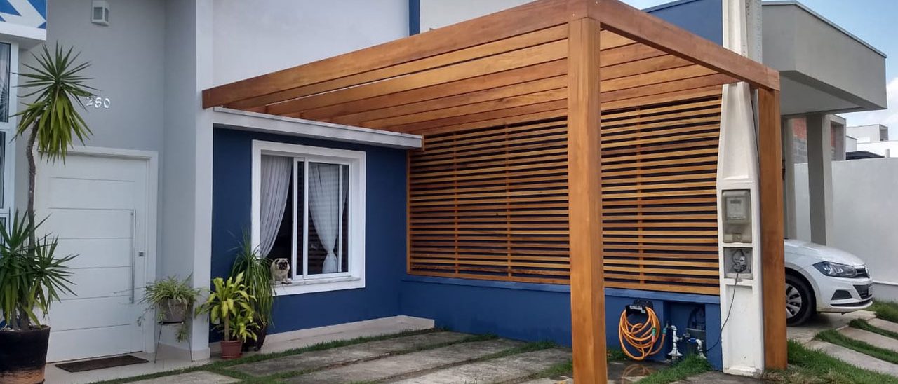 Pergolado de madeira serrada com ripas no teto e painel ripado lateral, na entrada de uma casa, servindo de garagem